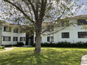 Wide shot of the courtyard-facing buildings with unit numbers partially visible, emphasizing the lush lawn, tree coverage, and modernized entryways for a welcoming environment.